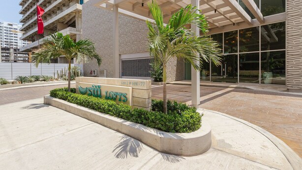 Entrada moderna de un edificio con un letrero que dice "SITI LOFTS" y un área exterior cubierta con palmeras.