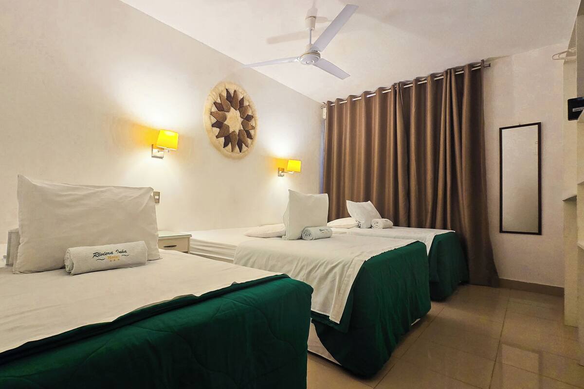A hotel room with two beds, each with a green bedspread, white pillows, and a rolled towel with text. There are two wall-mounted lamps, a ceiling fan, and a large window with curtains.