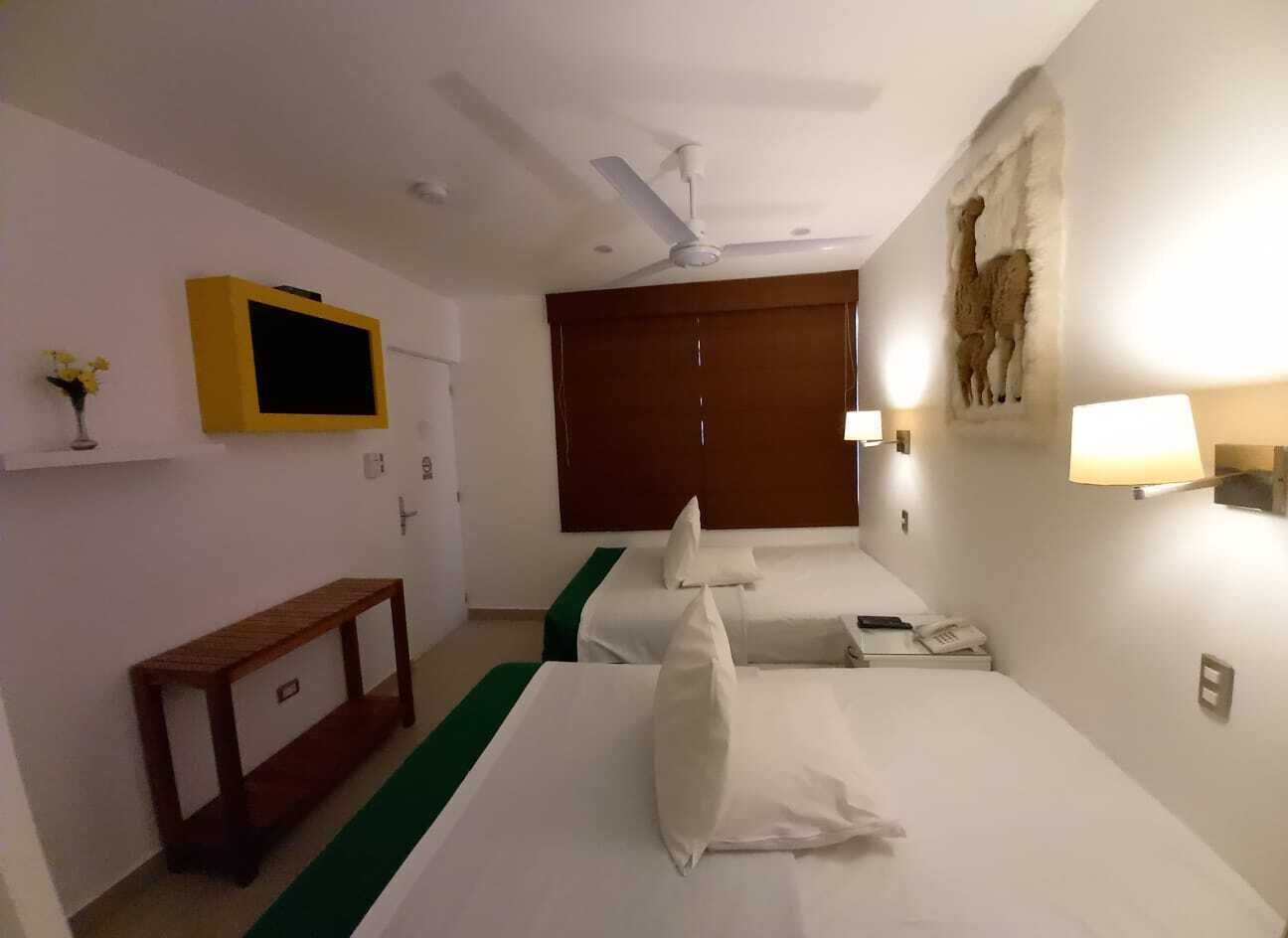 Una habitación de hotel con dos camas, televisión, escritorio y lámpara de pared.