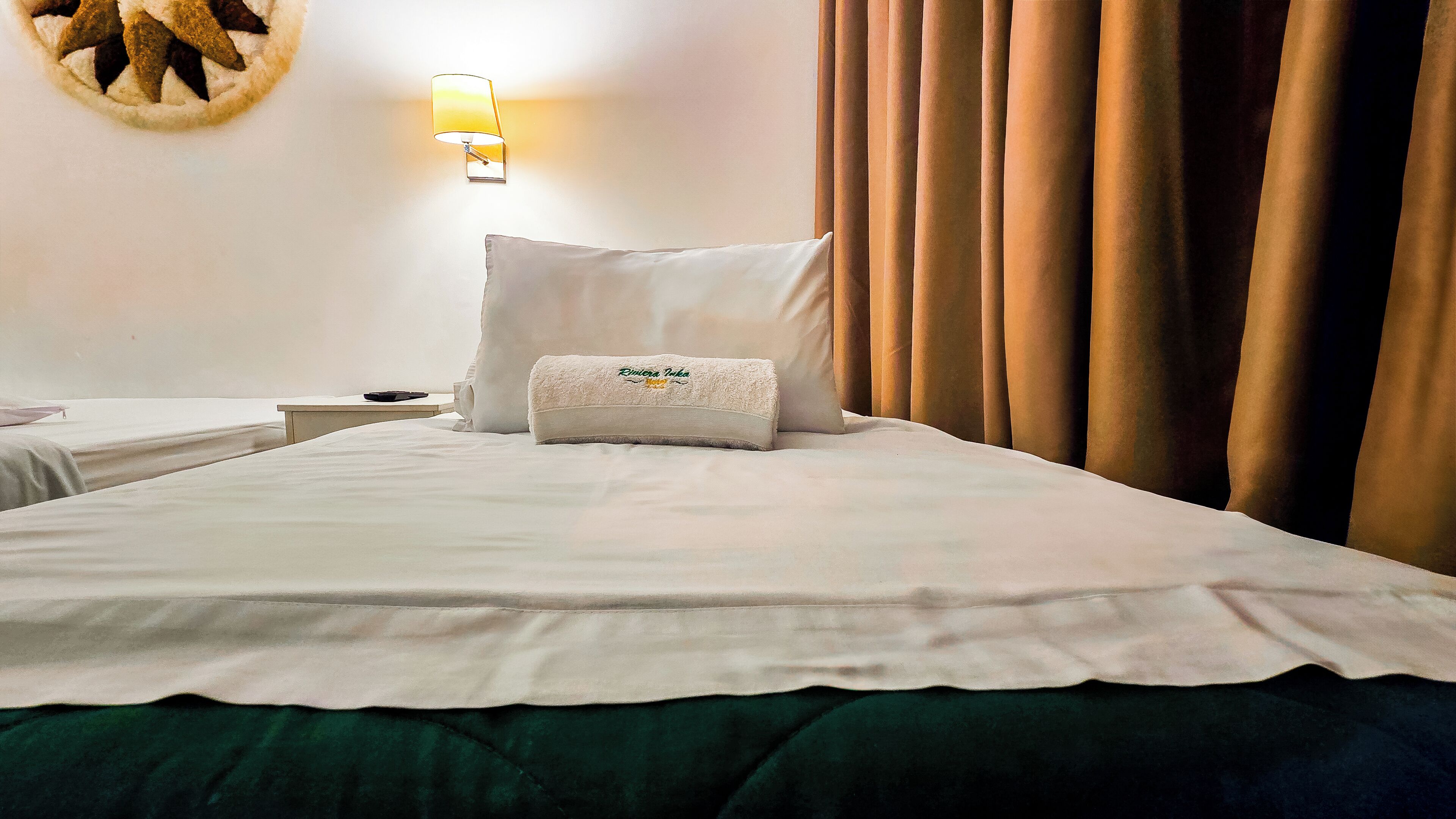 A neatly made bed with white linens, a pillow with a logo, a wall-mounted lamp, and a curtain.