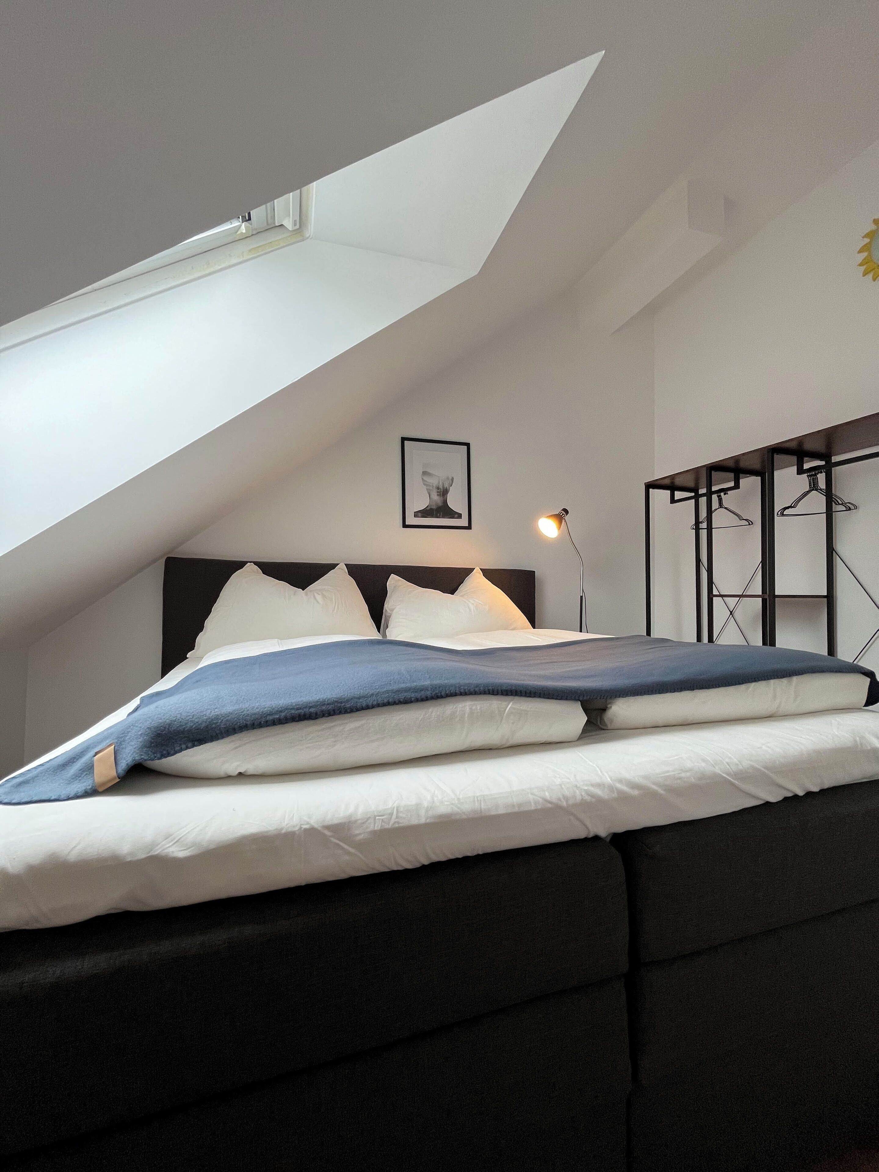 A bedroom with a sloped ceiling, a bed with white and blue bedding, a bedside lamp, a framed picture on the wall, and a black metal hanger rack.