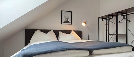 A bedroom with a sloped ceiling, a bed with white and blue bedding, a bedside lamp, a framed picture on the wall, and a black metal hanger rack.