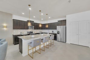 Une cuisine moderne avec des meubles de couleur sombre, un îlot central doté de tabourets de bar, des appareils en acier inoxydable et un éclairage suspendu.