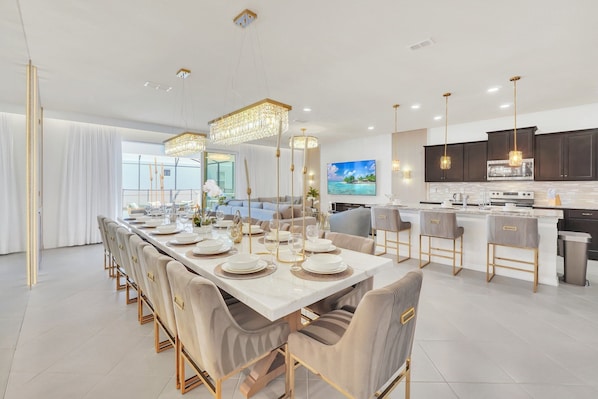 Un espace repas moderne avec une longue table dressée pour un repas, une cuisine aux meubles sombres et une grande fenêtre offrant une vue dégagée.