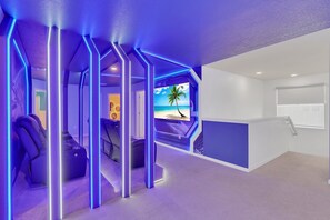 A modern room with a view of the beach, a massage chair, and a TV screen displaying a beach scene. - 2 Slides • Star Wars Theater • Mario Kart Game Room • Princess Castle (Clermont)
