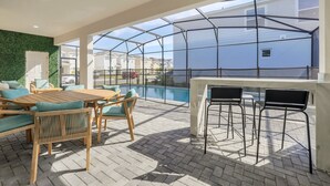 Une terrasse sur le toit avec une table et des chaises en bois, un bar fermé par des parois de verre, et une vue sur une piscine et les maisons voisines.