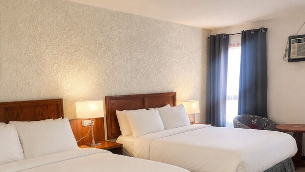 A hotel room with two beds, a nightstand, a lamp, a chair, and a window with curtains. - Maple Creek Motor Inn (Maple Creek)
