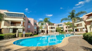 Piscina cercada por edifícios modernos com varandas, palmeiras e céu claro.