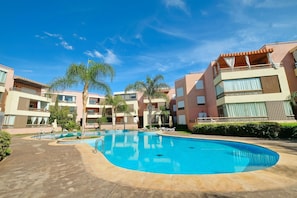 A swimming pool surrounded by multi-story buildings with balconies, palm trees, and a clear sky.
