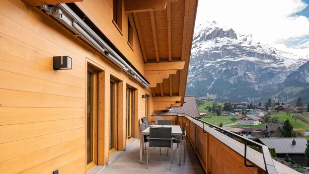 A wooden balcony with a table and chairs, overlooking a mountain and a village.