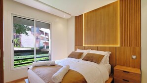 A modern bedroom with a large bed, wooden headboard, and a view of a building complex outside.