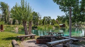 A lakeside campsite with picnic tables, a fire pit, and a cabin in the background.