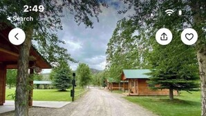 A rural road leading to a wooden cabin with a green roof, surrounded by trees and a grassy area.