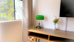Una habitación con un escritorio de madera, una lámpara de escritorio verde, un jarrón blanco con flores, un televisor montado en la pared y una ventana con cortinas.
