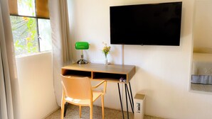 Una habitación con un escritorio de madera, una lámpara de escritorio verde, un televisor negro montado en la pared, una silla de mimbre y un pequeño cubo de basura.