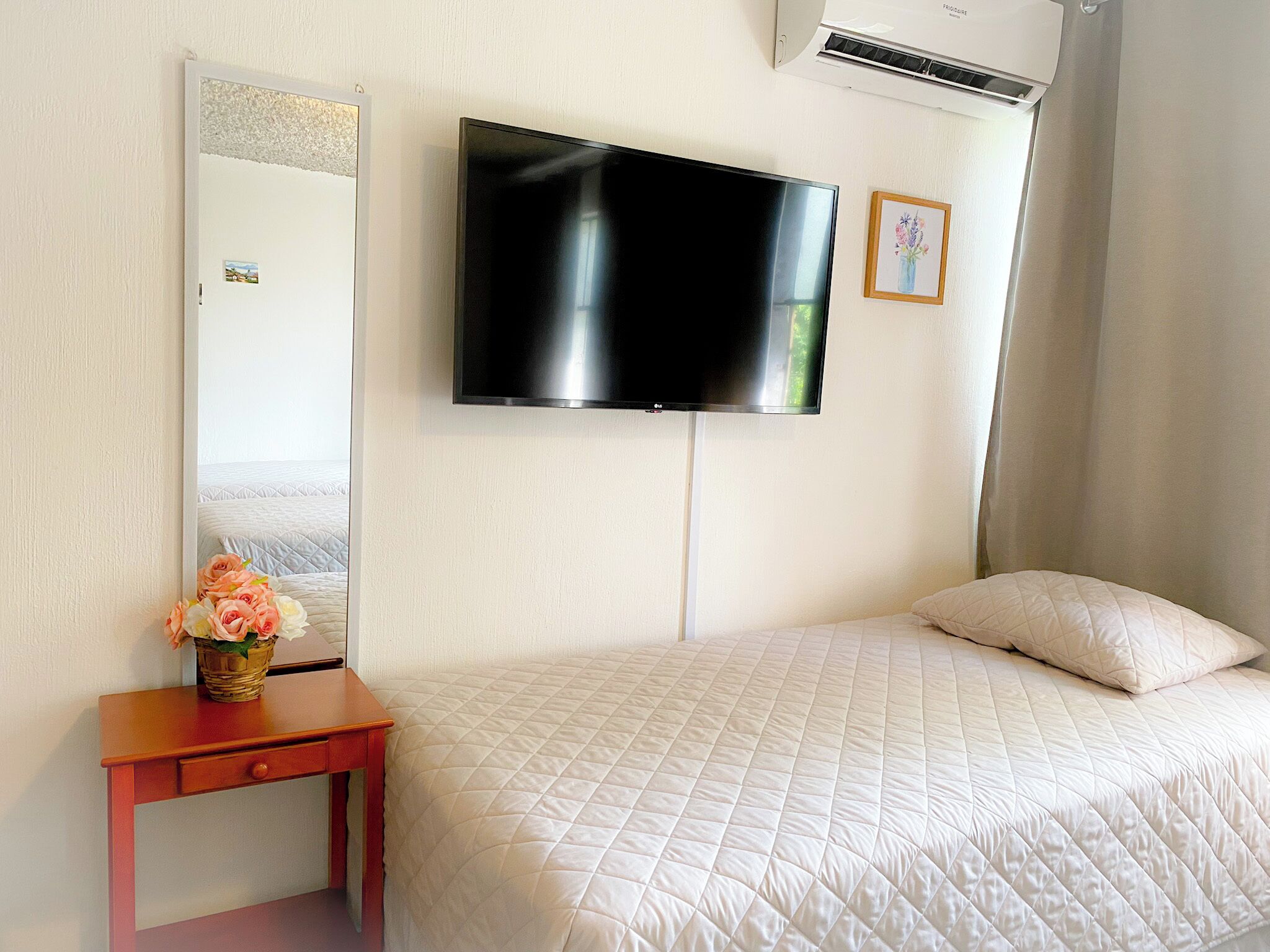 A bedroom with a bed, a TV mounted on the wall, a mirror, a small table with a vase of flowers, and an air conditioning unit.