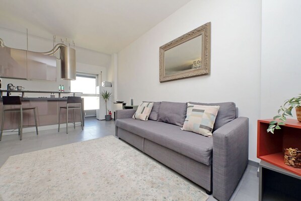 A modern living room with a grey sofa, a patterned rug, a kitchen in the background, and a large mirror on the wall. - Rome, Esquilino, apartment for 4 (Roma)