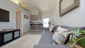 A modern living room with a grey sofa, a flat-screen TV mounted on the wall, a kitchen area in the background, and a large mirror.