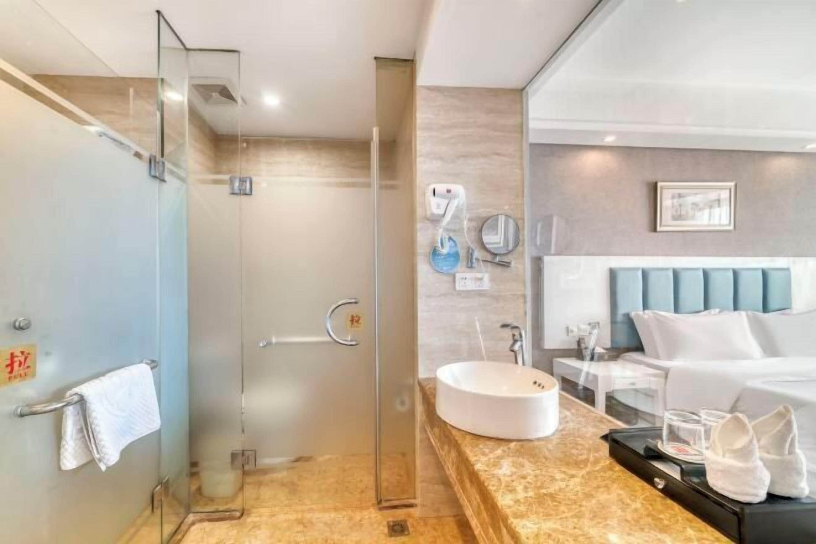 A modern hotel room with a glass-enclosed shower, a round sink, a bed with a blue headboard, and a bedside table with a lamp.