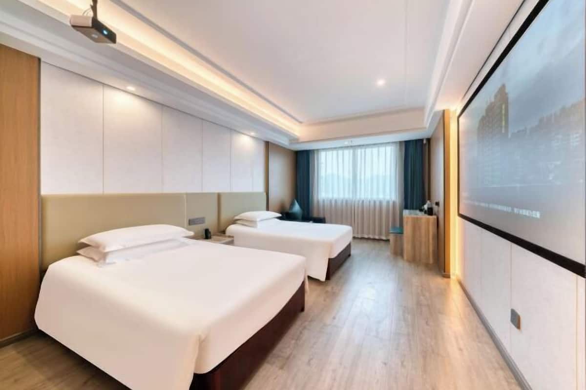 Une chambre d’hôtel avec deux lits, un bureau, une grande fenêtre avec des rideaux et un écran de projection fixé au mur.