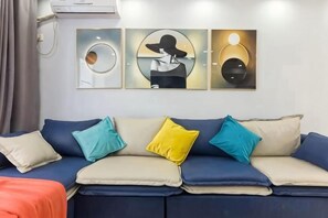 Een kamer met een blauwe bank, beige en blauwe kussens, en drie aan de muur hangende kunstwerken.