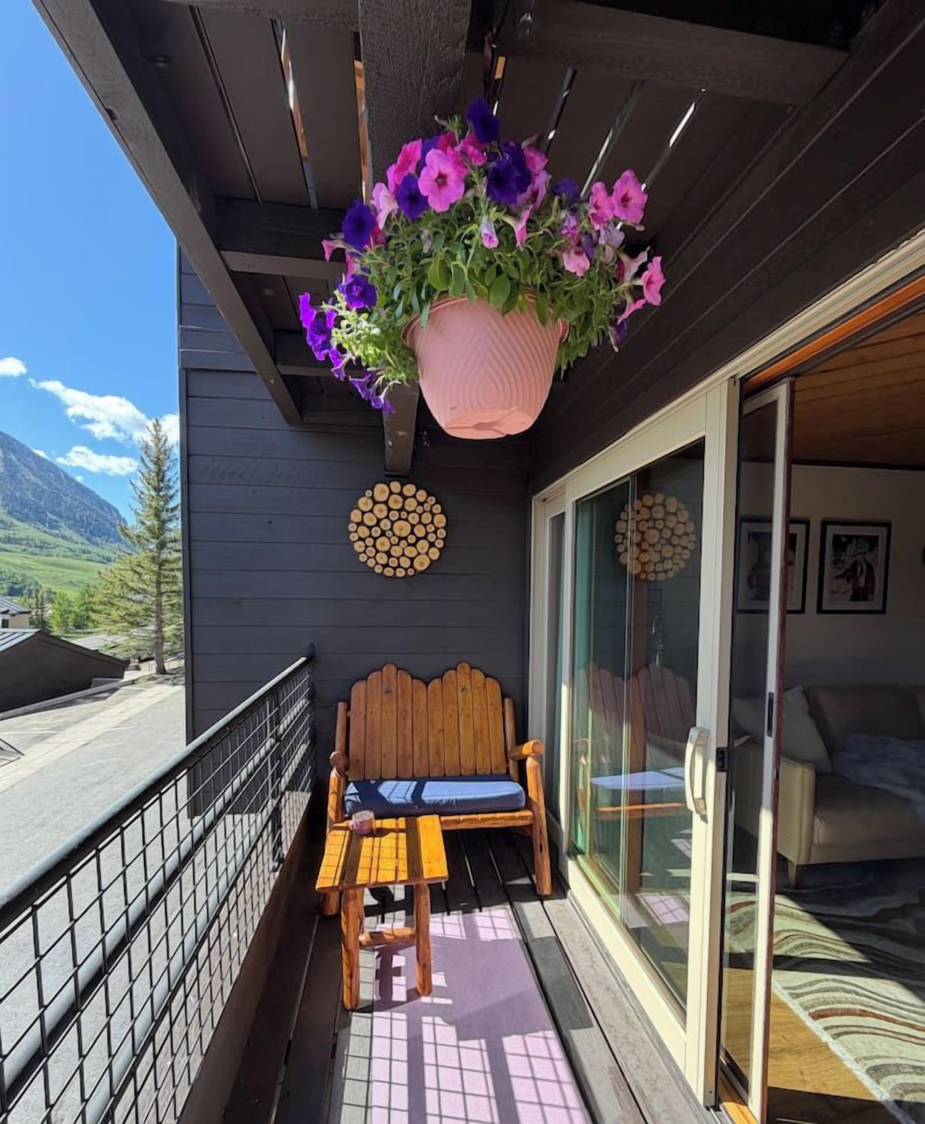 A balcony with a wooden bench and a table, a hanging flower pot, and a sliding glass door leading to a room with a bed.