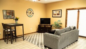 A living room with a gray sofa, a TV on a stand, a dining table with chairs, a clock, and a door with blinds. - Cozy 1-bedroom apartment in quaint Creedmoor with AC, WiFi &laundry (Creedmoor)