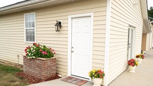 A beige single-story house with a white front door, a small window, and a brick planter with flowers. There are two potted plants on the sidewalk. - Cozy 1-bedroom apartment in quaint Creedmoor with AC, WiFi &laundry (Creedmoor)