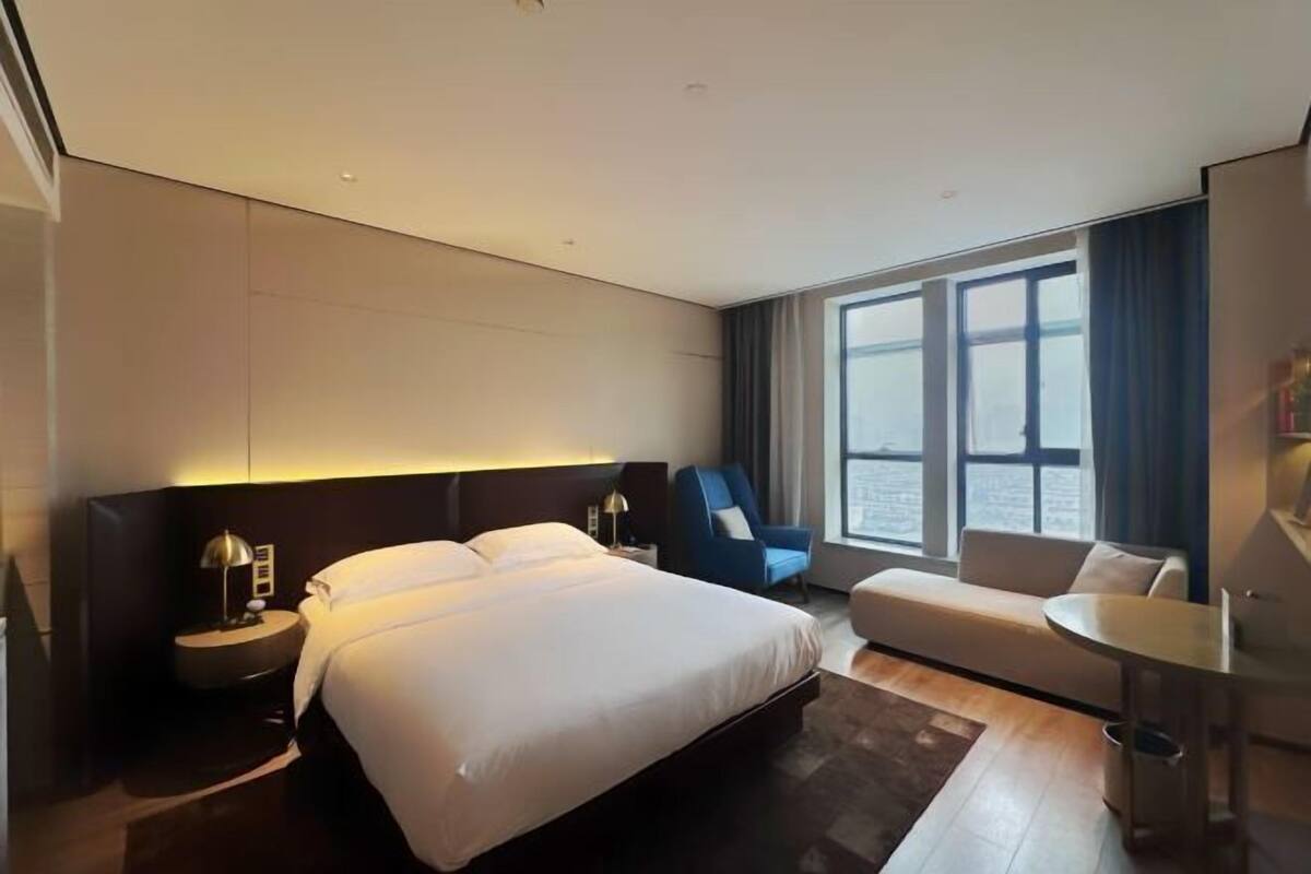 Une chambre d’hôtel moderne dotée d’un grand lit, d’un coin salon, d’une petite table et offrant une vue sur la ville grâce à de grandes fenêtres.