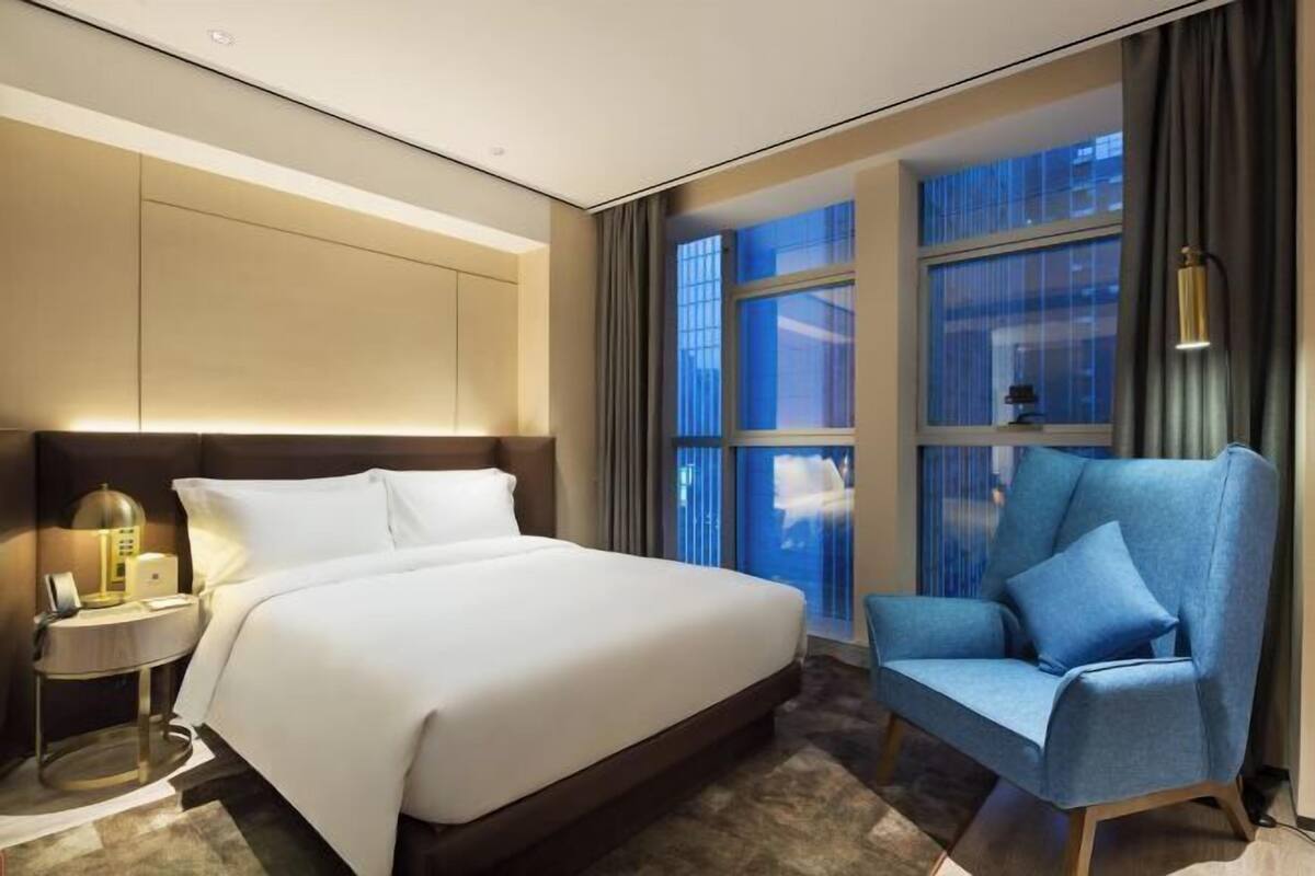 Une chambre d’hôtel avec un grand lit, une table de chevet avec une lampe, un fauteuil bleu et une vue sur la ville grâce à de grandes fenêtres.
