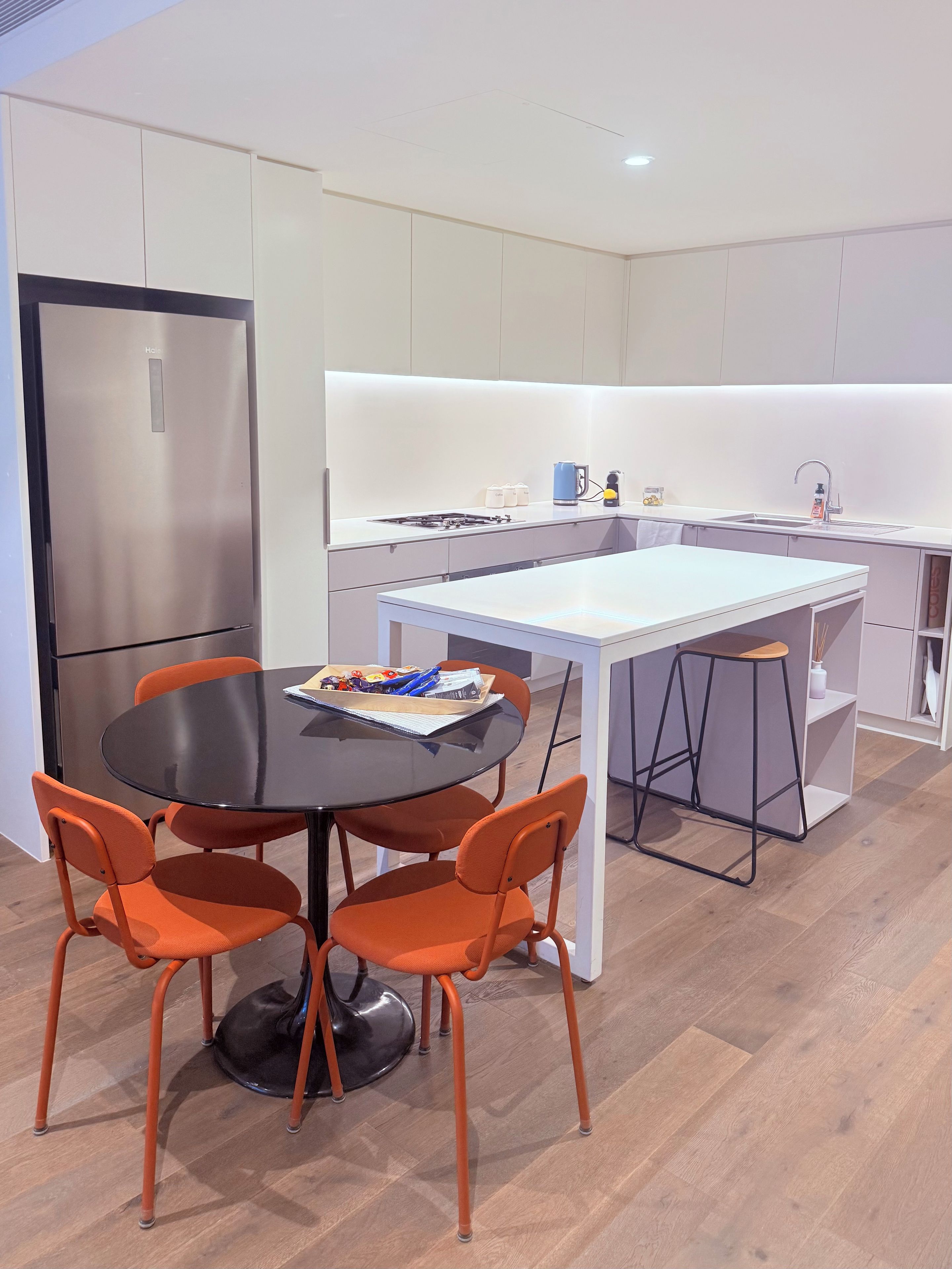 A modern kitchen with a stainless steel refrigerator, a white countertop, and a round dining table with orange chairs.