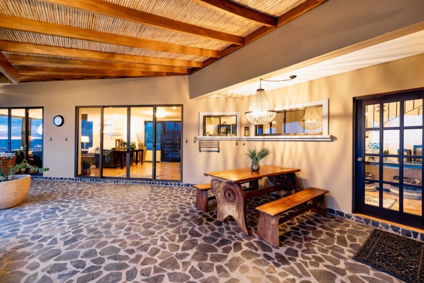 A spacious patio with a stone floor, a wooden table with benches, and a view of the ocean through large sliding doors.