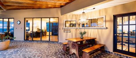A spacious patio with a stone floor, a wooden table with benches, and a view of the ocean through large sliding doors.