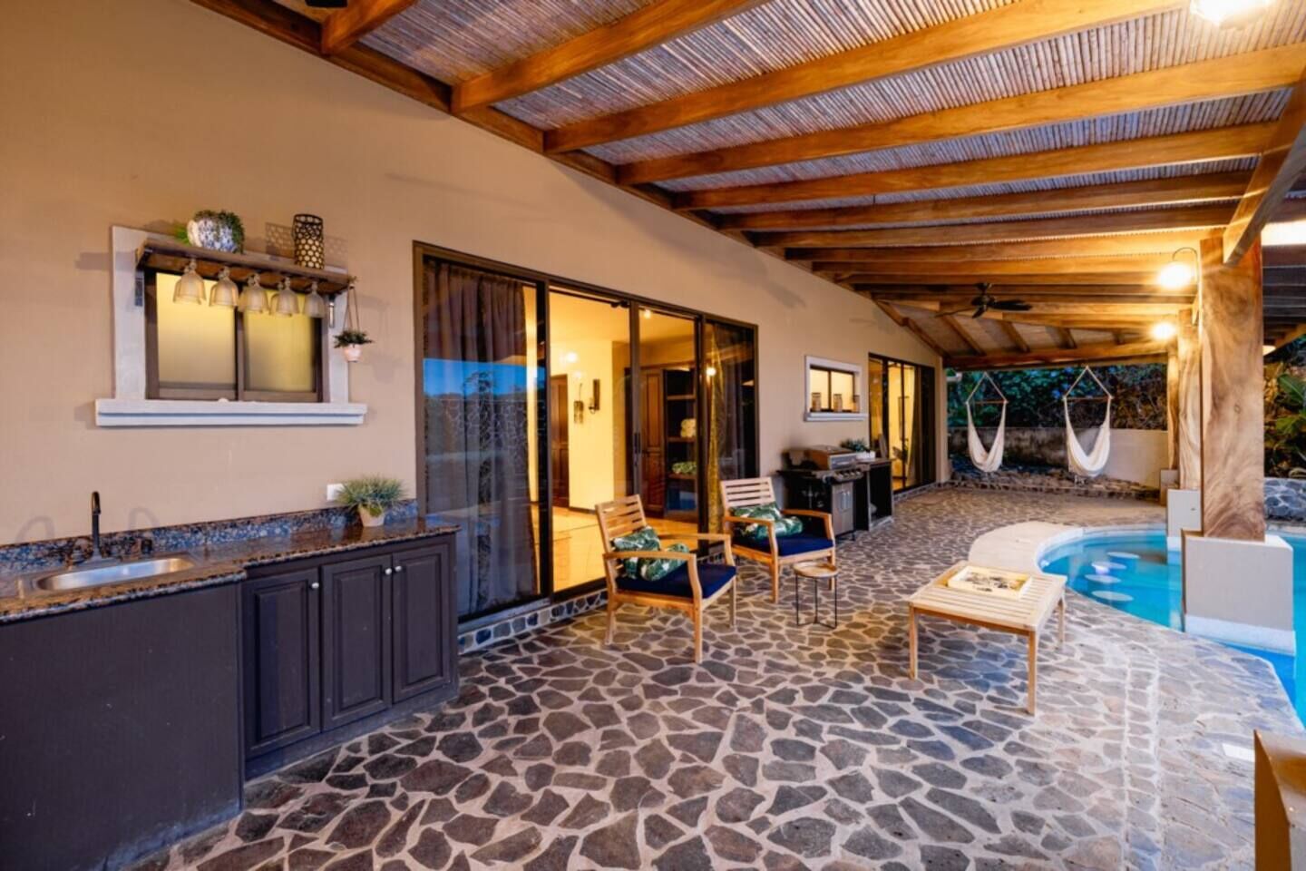 A covered outdoor area with a stone floor, a pool, and a dining setup.