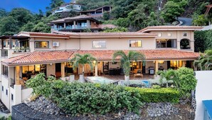 Una casa de varios pisos con piscina, rodeada de abundante vegetación y un paisaje montañoso.