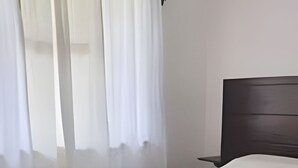 Uma cama bem arrumada com colcha e toalha brancas, cabeceira escura e janela com cortinas brancas e leves.