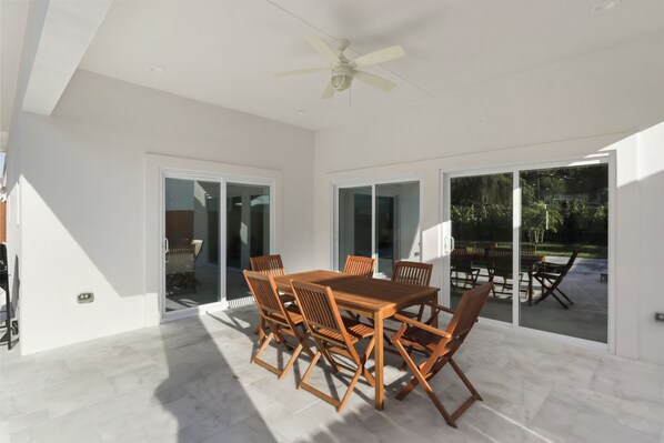 A spacious patio with a wooden table and chairs, large sliding glass doors leading to an outdoor area, and a ceiling fan.