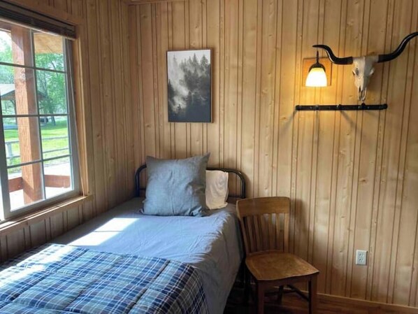 A wooden-paneled room with a bed, a chair, and a wall-mounted light fixture with a cow skull. - Longhorn Cabin At Mountain River Ranch (Ririe)