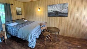 Une pièce avec un lit, une chaise, une petite table, des murs en bois et un tableau représentant une vache.
