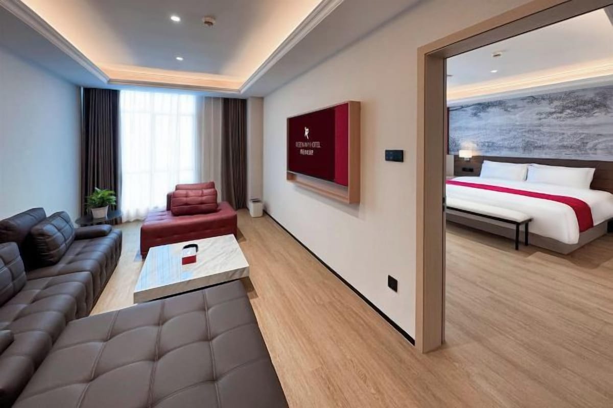 A modern hotel room with a sofa, a coffee table, a bed with a red accent, and a large window.