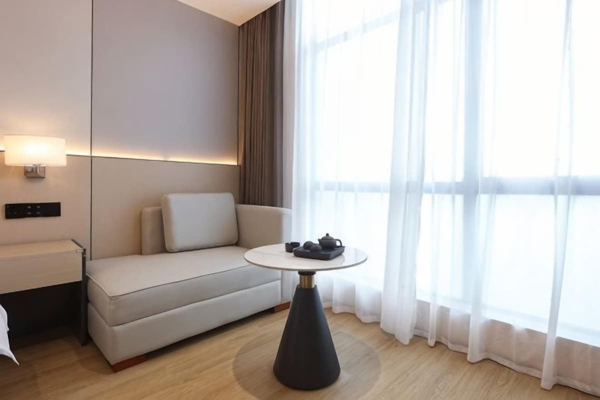 A modern hotel room with a sofa, a small round table with a teapot, and a large window with sheer curtains.