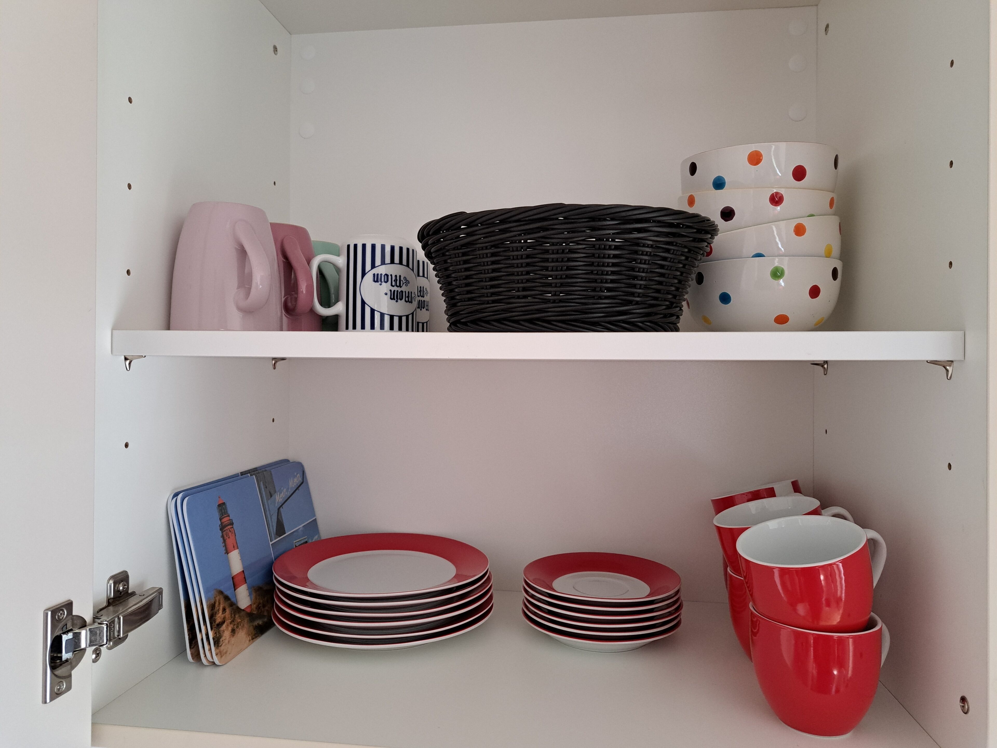 Une cuisine avec des meubles blancs, un étendoir suspendu où sont accrochées des assiettes et des tasses, et un panier posé sur l’étagère.