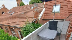 An outdoor balcony with two grey cushioned chairs and a table, a blue umbrella, and a view of a red-tiled building with a chimney. - A millside dream, stylish, right next to the historic mill, not far from the harbor... (Ditzum)