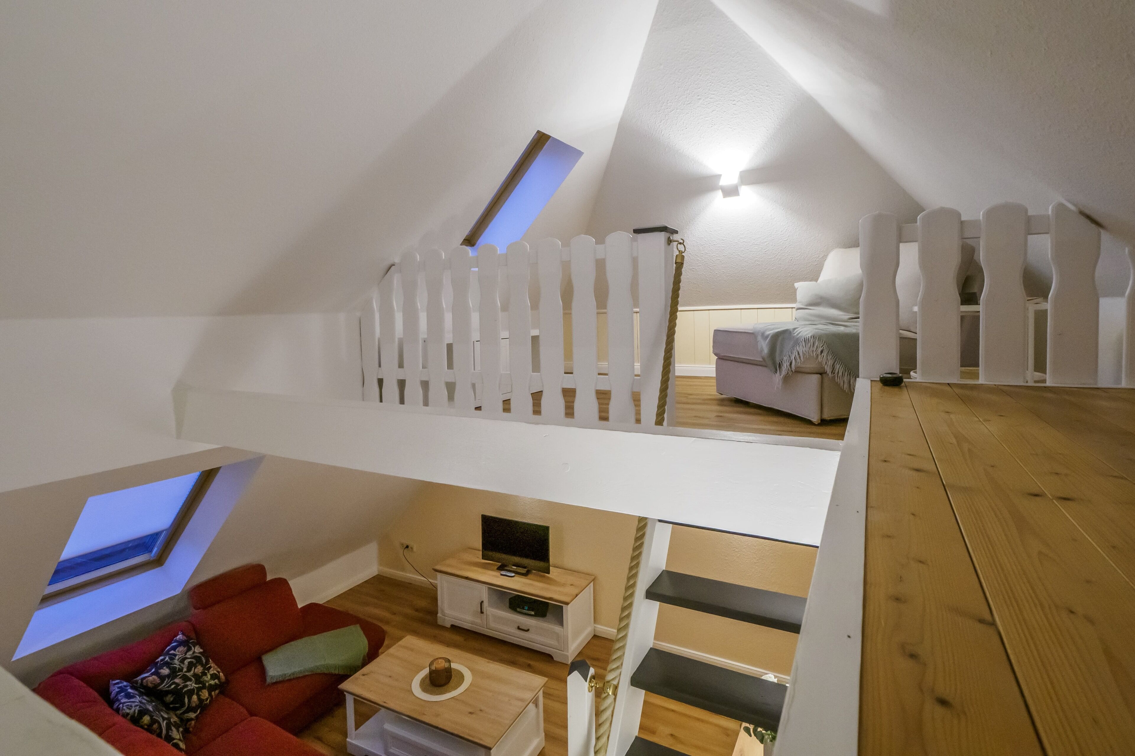 An attic bedroom with a sloped ceiling, a white picket fence, a red sofa, a small TV stand, and a wooden floor.