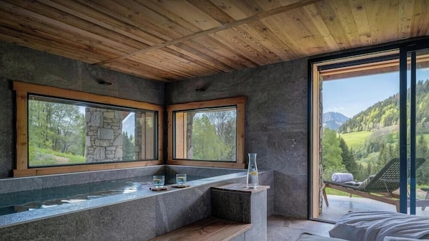 A modern bathroom with stone walls, wooden ceiling, and large windows offering a view of a green landscape.