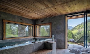 Un bagno moderno con pareti in pietra, soffitto in legno e ampie finestre che si affacciano su un paesaggio verde.