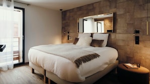 A modern bedroom with a large bed, a wooden headboard, a mirror, and a small side table with a book and glasses.