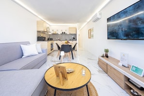 Un soggiorno moderno con un divano, un tavolino rotondo, un'area pranzo con sedie, una cucina sullo sfondo e un grande televisore montato a parete.