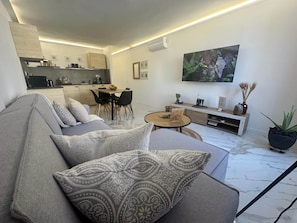 Un soggiorno moderno con un divano grigio, un'area pranzo, una televisione a schermo piatto e una cucina sullo sfondo.