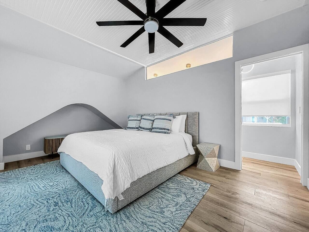 A modern bedroom with a bed, a ceiling fan, a skylight, and a view of the outdoors.